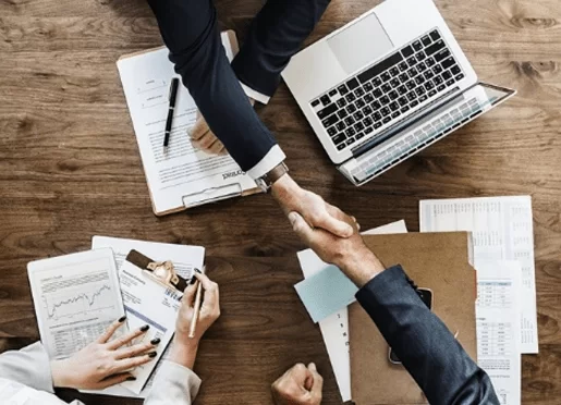 Executivos apertando as mãos durante uma negociação de contrato em uma mesa com documentos e laptop.